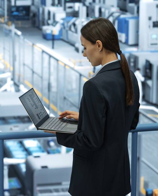 Portrait of a Caucasian Female Engineer Using Laptop Computer and Looking Around a Factory Facility with Robotic Arms Producing Modern Electronic Components for Tech Industries